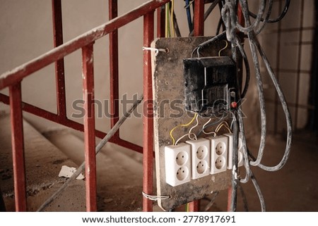 Makeshift temporary electrical panel with sockets and exposed wiring attached to a rusty metal railing on a concrete staircase at a construction site.
