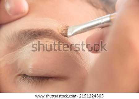 A makeup artist adding concealer on the top of a young woman's eyebrows with a thin angled brush. Shaping technique and avoiding plucking.
