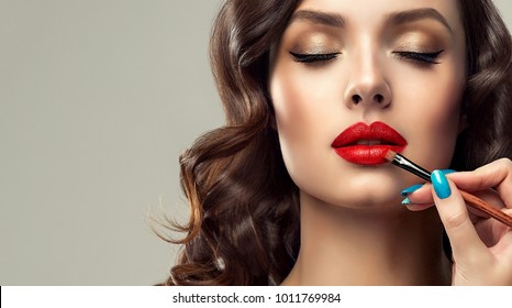  Makeup artist applies  red lipstick  . Beautiful woman face. Hand of make-up master, painting lips of young beauty  model girl . Make up in process