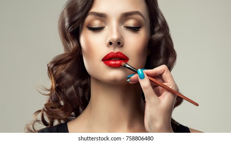  Makeup artist applies  red lipstick  . Beautiful woman face. Hand of make-up master, painting lips of young beauty  model girl . Make up in process
