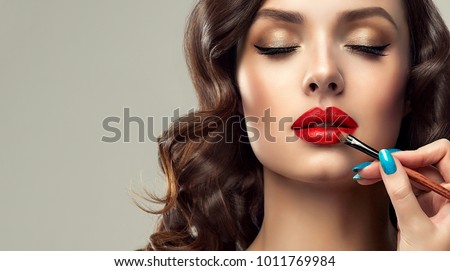  Makeup artist applies  red lipstick  . Beautiful woman face. Hand of make-up master, painting lips of young beauty  model girl . Make up in process