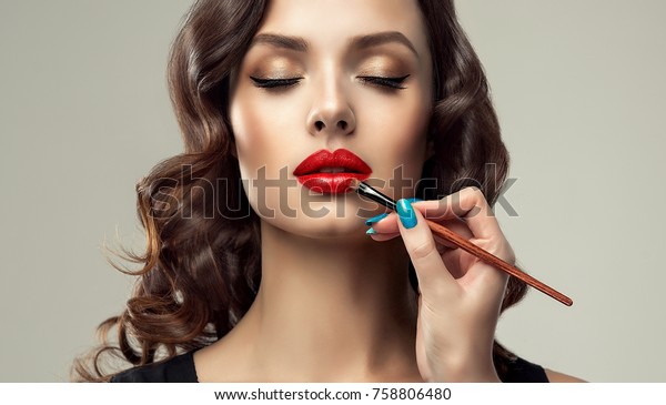 メイクアップアーティストは赤い口紅を塗ります 美しい女性の顔 手作り師匠の手描きの若い美容師の唇 メークアップの処理中 の写真素材 今すぐ編集