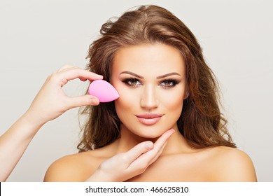 Makeup artist applying foundation on gorgeous young woman's face using pink beauty sponge. Skin foundation blender application by make-up artist. Closeup, studio lighting, retouched.