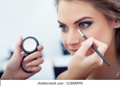 Make-up artist applying white eyeshadow in the corner of model's eye and holding a shell with eyeshadow on background, close up