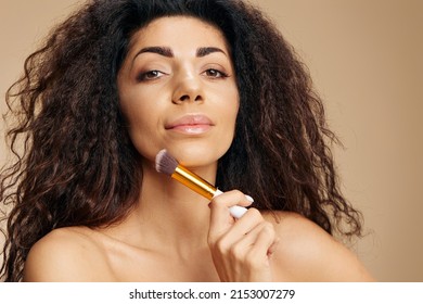 MAKEUP ARTIST CONCEPT. Enjoyed smiling tanned awesome curly Latin lady hold makeup brush after makeup visage ready to party posing isolated on pastel beige background. Closeup banner cosmetic offer