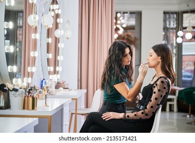 Makeup artist doing professional makeup in visage studio. Stylish woman sitting at dressing table while female beauty specialist in sterile gloves applying lipstick on client lips with cosmetic brush.