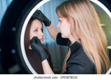 Makeup Artist Holds A Thin Coloring Thread Against The Face Of A Client, Marking An Outline Of Brows, All In Frame Of Ring Light Lamp. Permanent Make Up And Brow Making Professional Sphere.