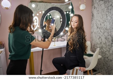 Makeup artist photographing client in beauty salon. Young woman with long hair sitting on chair and looking at beauty specialist while female worker taking picture with smartphone.