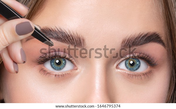 Make-up\
artist plucks eyebrows with tweezers to a woman with curly brown\
hair and nude make-up. Beautiful thick eyebrows close up.\
Professional makeup and cosmetology skin\
care.