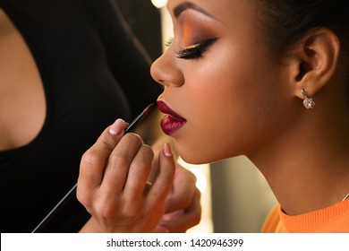 Makeup Artist Working With African American Model In Beauty Salon. Perfect Skin Facial. Beautiful Fashion Luxury Makeup Orange-coloured Shades. Big Lush Beautiful Lips. Sexy Violet Purple Pucker.