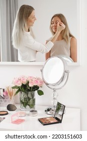 Makeup artist working with model reflected in mirror Cosmetics for make-up and roses flowers on table. Decorative cosmetics, brushes, eyes shadow with foundation on dressing table in beauty salon.