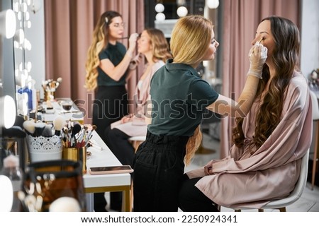 Makeup artists doing professional makeup for women in visage studio. Young woman beauty specialist in sterile gloves applying eyeshadow on client eyelid with cosmetic brush.