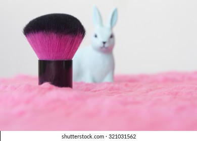 Makeup brush and rabbit plastic toy on pink fluffy cloth. Blur image, selective focus on the brush. Perfect as background for girls and makeup artists. Empty space for copywriting.