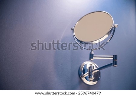 A makeup mirror is hanging on a wall. The mirror is silver and has a circular shape. The wall is blue.