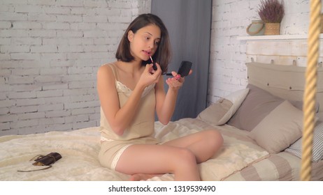 Makeup in the morning at home. Beautiful young woman sitting on bed put lipstick. Attractive caucasian lady holding little mirror wearing in pajamas in flat.