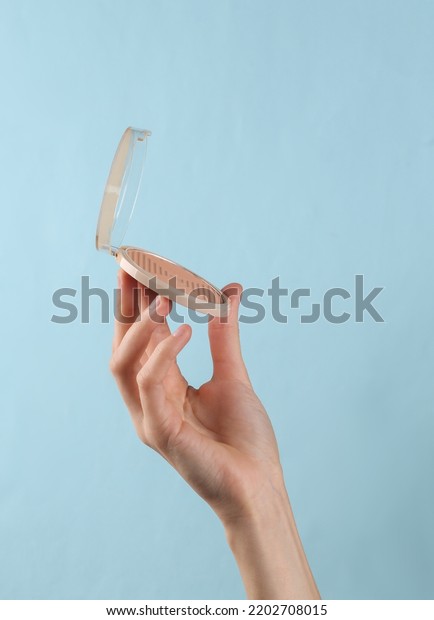 Makeup\
powder box in female hand on blue\
background.