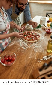 Making The Best Pizza. Young Couple Making Pizza Together At Home. Man In Apron Professional Cook Adding Basil On The Dough While Woman Helping Him. Selective Focus On Pizza. High Angle