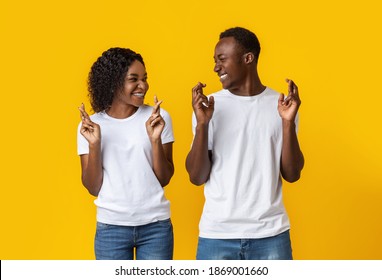 Making Cherished Wish. Young Cheerful African-american Couple With Crossed Fingers Looking At Each Other And Smiling, Yellow Studio Background. Emotional Black Man And Woman Praying For Good