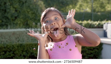 Making child in pink top pulling cheeks and sticking tongue in sunlit backyard, with emoji icons. Kid, garden, playful, cheerful, sunny, outdoors, candid