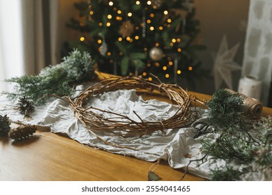 Making Christmas rustic wreath. Evergreen branches, pine cones, twigs, twine, scissors on rustic table on backround of christmas tree lights. Winter holiday preparations - Powered by Shutterstock