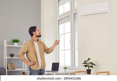 Making Domestic Life Easy And Enjoying Good And Convenient Conditioning System: Young Man Standing In Living-room, Holding Air Conditioner Remote Control And Adjusting Temperature At Home