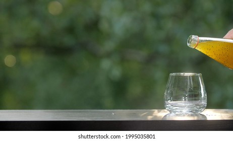 Making drinks outdoors. Drink bottle is tilted towards the empty glass. Orange drink with fruit pieces. Brown wooden table with green morning summer foliage bokeh background. Sun's rays glare