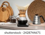 Making drip coffee. Glass chemex coffeemaker with paper filter, jar of beans and kettle on wooden countertop in kitchen