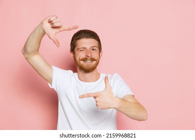 Making Finger Frame. Half-length Portrait Of Young Happy Man In White T-shirt Posing Isolated On Pink Background. Concept Of Art, Fashion, Emotions, Aspiration. Copy Space For Ad