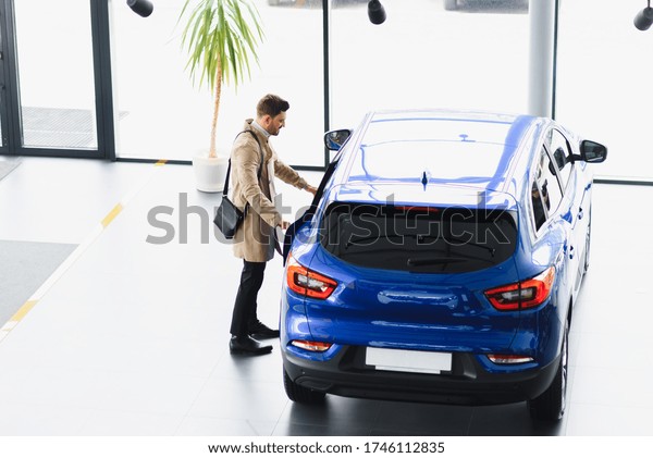 Making his\
choice. Horizontal portrait of a young man in a suit looking at the\
car and thinking if he should buy\
it