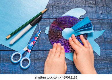 Making homemade toy fish from the CD. Paper, pencil, marker glue gun on blue wooden table. Handmade children's project. Step by step photo instructions. Step 6: The child glues details toys