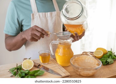 Making kombucha tea drink at home, fermented drink flavoured with ginger, lemon and mint. Healthy drink, organic probiotic beverage. Black young male hands pouring drink through a sieve. - Powered by Shutterstock