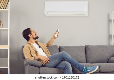 Making Life Easy And Enjoying Convenient Conditioning System At Home. Happy Relaxed Young Man Sitting On Sofa In Living-room, Turning On Air Conditioner And Regulating Temperature With Remote Control