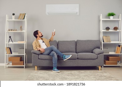 Making Life Easy And Enjoying Good, Modern, Convenient Conditioning System At Home: Relaxed Young Man Sitting On Sofa In Living-room And Adjusting Air Conditioner Temperature Mode With Remote Control