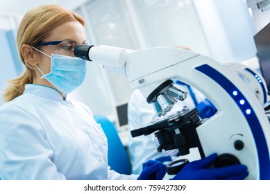 Making Medical Discovery. Serious Blond Young Researcher Wearing A Uniform And A Medical Face Mask And Gloves And Glasses And Looking In The Microscope While Being In The Lab