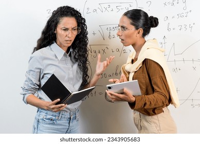 Making mistakes and wrong answer concept. Two young female students of Oriental appearance cannot solve a mathematical equation, they are puzzled, they hold a textbook and a tablet in their hands