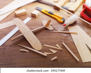 Making Model Airplane From Wood. Wooden Air Plane Handcrafted With Balsa Wood, On Work Table By The Window. Airplane, Cutter Knife, Balsa Wood Material And Glue On Table.