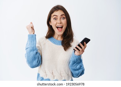 Making Money In Internet. Excited Woman Raising Hand Up And Shouting Yes After Winning Online, Achieve Goal In App, Holding Smartphone, White Background.