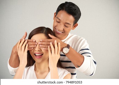 Making Pleasant Surprise To Girlfriend: Handsome Asian Man Standing Behind Young Woman And Covering Her Eyes With Hands, Studio Shot