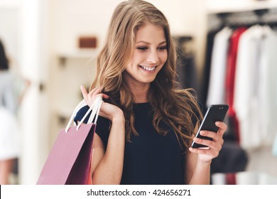 Making The Retail Connection. Beautiful Young Woman With Shopping Bags Using Her Smart Phone With Smile While Standing At The Store