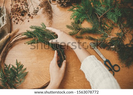 Making rustic christmas wreath flat lay. Hands holding fir branches and pine cones, thread, scissors on wooden table. Details for workshop of making christmas wreath