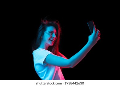 Making selfie. Half-length portrait of young beautiful girl with smartphone isolated on dark background in neon light. Concept of human emotion, facial expressions, youth culture. Copy space for ad