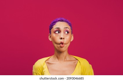 Making a silly face. Quirky young woman making a funny face while standing against a pink background. Fashionable young woman wearing makeup with purple hair in a studio.