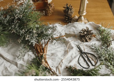 Making stylish Christmas rustic wreath. Evergreen branches, pine cones, twigs, twine, scissors on rustic table in atmospheric festive room. Winter holiday preparations, details - Powered by Shutterstock