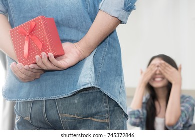 Making A Surprise. Boyfriend Hiding Red Gift Box Behind His Back While His Girlfriend Is Covering Her Eyes With Hands And Sitting On Couch At Home.