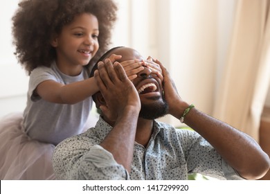 Making surprise for daddy concept, cute little african american kid daughter close eyes of excited happy black dad on fathers day, funny small child girl having fun with papa laughing bonding at home