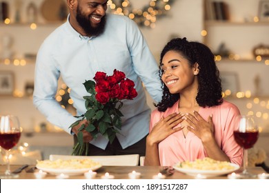 Making Surprise. Excited beautiful african american girlfriend receiving bouquet of red roses from her happy boyfriend who holding bunch of flowers during romantic dinner in luxury restaurant or cafe