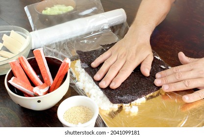 Making Sushi At Home. Close-up Of A Young Caucasian's Hand Pressing A Nori Leaf Against Rice On A Board With Cling Film. Rolls With Crab Sticks, Cheese,cucumber. Steps To Make Salmon Sushi,DIY Sushi