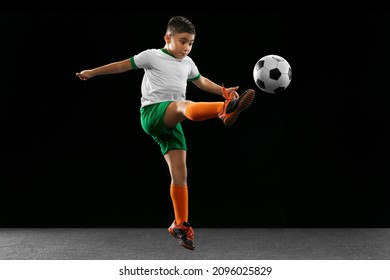 Making A Toe Punt. Portrait Of Boy, Child, Football Player In Uniform Training, Kicking Ball Isolated Over Black Background. Concept Of Action, Team Sport Game, Energy, Vitality. Copy Space For Ad.