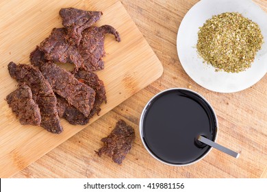 Making Traditional Homemade Beef Jerky Or Biltong With Thin Strips Of Lean Beef Meat On A Cutting Board Alongside Teriyaki Sauce And Dried Oregano, Overhead View