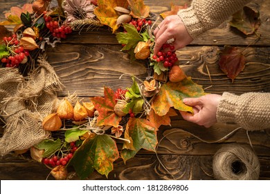 Making wreath autumn colorful leaves and natural materials on rustic wooden boards. Top view women's hands make round wreath autumn harvest and foliage on brown wooden table. Decoration for interior.